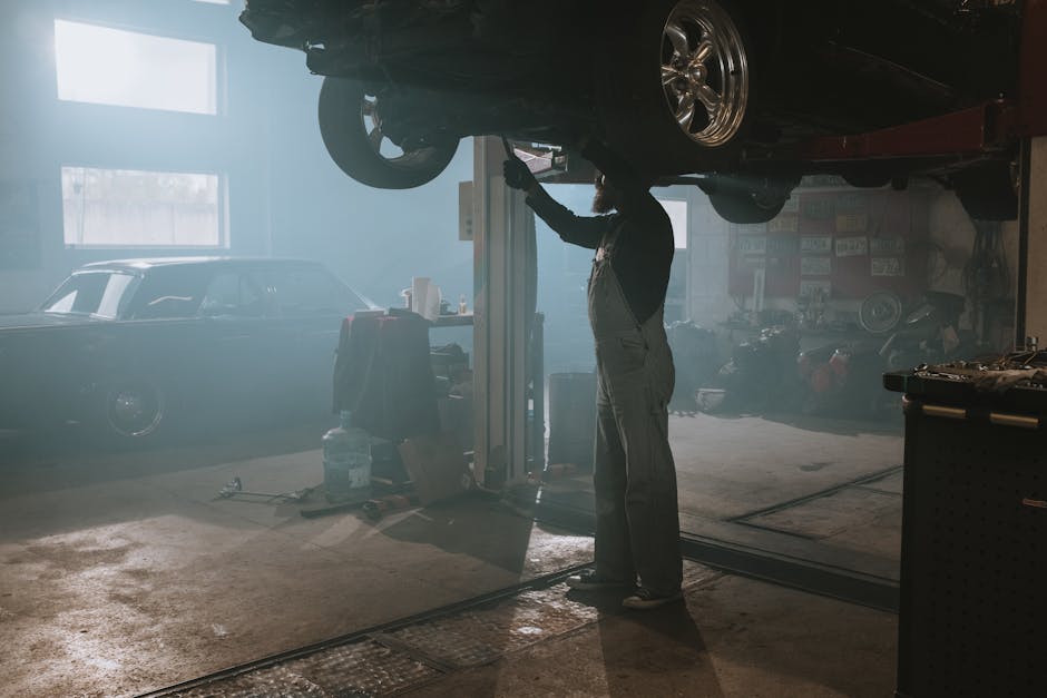 A mechanic is fixing a car raised on a lift in a dimly lit garage, creating a vintage ambiance.