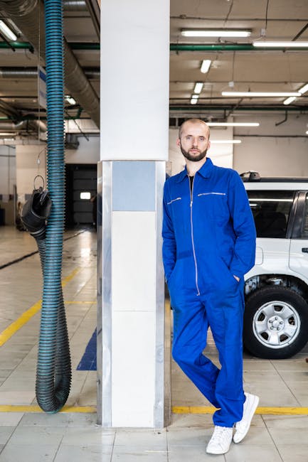 Bald bearded mechanic in blue coveralls leaning against a pillar in an auto workshop. Hands in pockets.