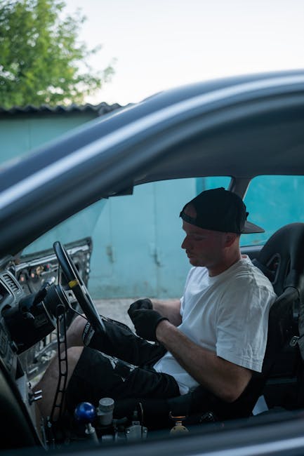 A mechanic in a black cap and gloves adjusts the steering wheel inside a car.