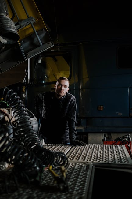 Mechanic posing in a dimly lit repair shop with a truck and tools around.